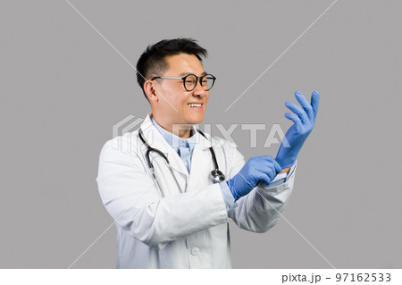 Smiling mature chinese man therapist in white coat, glasses puts on protective gloves on hands 97162533