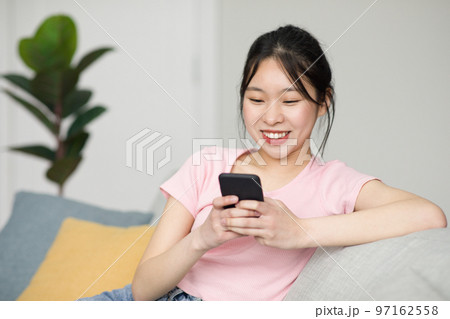 Excited korean lady sitting on sofa and using smartphone, chatting with friends online, checking social media Excited korean lady sitting on sofa and using smartphone, chatting with friends online, checking social media 97162558