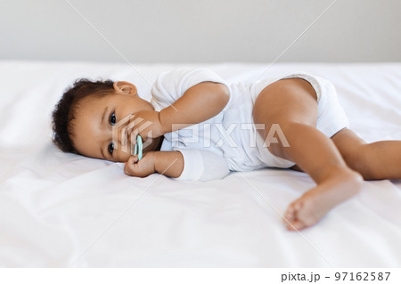 Teething Concept. Cute Little Black Baby Biting Teether While Lying In Bed Teething Concept. Cute Little Black Baby Biting Teether While Lying In Bed 97162587