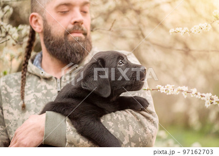 A small black Labrador retriever puppy. A dog in the arms of a bearded man. 97162703