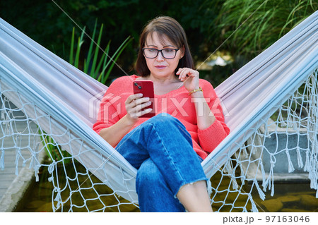 Middle aged woman with smartphone sitting in hammock 97163046