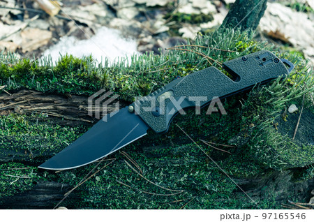 A sharp military knife with a black blade. Knife with a green handle on the moss in the forest. 97165546
