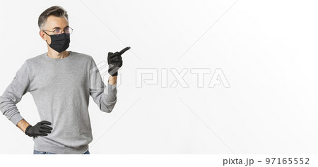 Concept of coronavirus, lifestyle and quarantine. Intrigued middle-aged man in medical mask, gloves and glasses pointing finger right and looking at advertisement, standing over white background 97165552