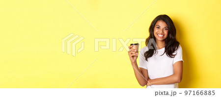 Portrait of attractive african-american woman in white t-shirt, drinking coffee and smiling, standing over yellow background 97166885