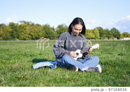Singing asian girl playing ukulele on grass, sitting on blanket in park, relaxing outdoors on sunny day 97168030