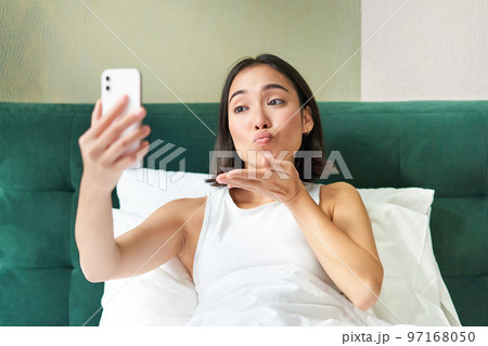 Beautiful asian girl lying in bed, making morning selfie, taking picture on smartphone in bedroom, smiling happily Beautiful asian girl lying in bed, making morning selfie, taking picture on smartphone in bedroom, smiling happily 97168050