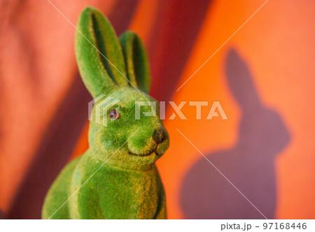 Green cute funny Easter bunny with long ears on the orange sunny background in springtime. Easter holiday in April. Toy decorative dreamy hare - a symbol of Easter. Smiling rabbit portrait close-up 97168446