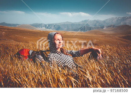 Tourist woman relax sitting in orange grass field. Mountain range in background. Autumn colorful scene. Backpack girl enjoy sunset light. Stunning nature scenery. Travel, adventure, concept image Tourist woman relax sitting in orange grass field. Mountain range in background. Autumn colorful scene. Backpack girl enjoy sunset light. Stunning nature scenery. Travel, adventure, concept image 97168500