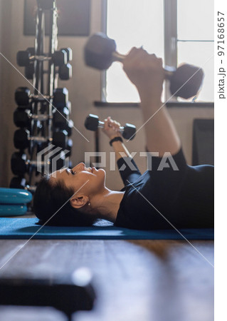 Top view portrait brunette sports woman lying on floor mat holding dumbbells exercising at gym Top view portrait brunette sports woman lying on floor mat holding dumbbells exercising at gym 97168657