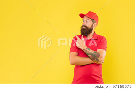 Serious deliveryman in casual red cap and tshirt pointing finger aside copy space, advert 97169679