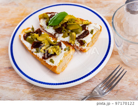 Tasty sandwich with cheese, sun-dried tomatoes, pesto sauce and olives on the white plate with blue line on the tablecloth 97170122
