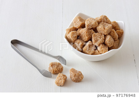 Cube-shaped cane sugar in wooden bowl on a white wooden table with silver sugar tongs.  High angle closeup, no people. 97176948