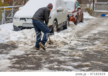 A man removes snow and ice stuck to the road with a shovel from the sidewalk. 97183929