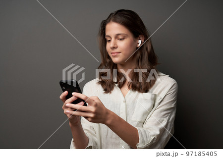Lovely casual teen girl in earbuds and smartphone in hands, standing on dark background 97185405