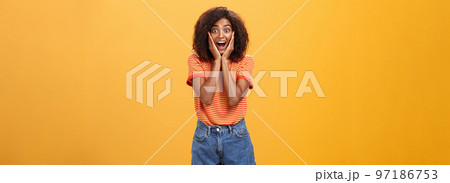 Impressed talkative african american woman finding out hot details of rumor being excited, thrilled leaning face on palms, smiling broadly with amused expression listening carefully over orange wall Impressed talkative african american woman finding out hot details of rumor being excited, thrilled leaning face on palms, smiling broadly with amused expression listening carefully over orange wall 97186753