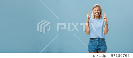Optimistic faithful good-looking young woman with blond hair and tattoos smiling joyfully crossing fingers for good luck waiting for dream come true making wish while standing over blue background Optimistic faithful good-looking young woman with blond hair and tattoos smiling joyfully crossing fingers for good luck waiting for dream come true making wish while standing over blue background 97186762