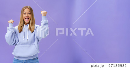 Cheerful happy and supportive young girlfriend with fair hair and tan in warm hoodide raising fists in cheer and triumph smiling saying yeah celebrating goal or success over purple background 97186982