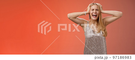 Upset distressed pressured fed up panicking young blond woman in dress scream cry heart out being heartbroken close eyes hold hands head mentaly unstable standing sad depressed red background 97186983