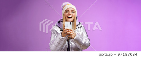 Amused excited attractive blond girlfriend holding smartphone up recording video boyfriend step snowboard first time capturing memories mobile camera standing happily purple background 97187034