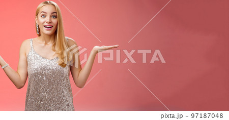 Carefree fascinated young blond girl 20s in silver glittering evening party dress raising hands sideways weighing shrugging smiling amused search right choice, deciding what do red background Carefree fascinated young blond girl 20s in silver glittering evening party dress raising hands sideways weighing shrugging smiling amused search right choice, deciding what do red background 97187048