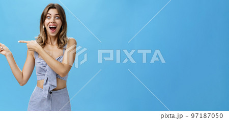 Unaltered shot of excited amused adult attractive woman with gapped teeth pointing left and smiling broadly from happiness and joy standing delighted and thrilled over blue background Unaltered shot of excited amused adult attractive woman with gapped teeth pointing left and smiling broadly from happiness and joy standing delighted and thrilled over blue background 97187050
