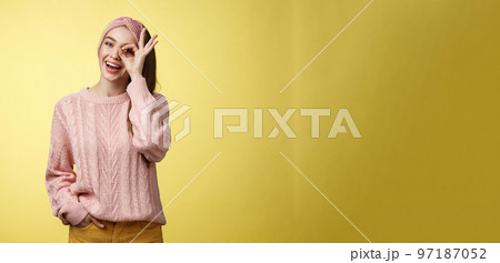 Cheerful girl saying ok. Attractive stylish shoolgirl in knitted sweater relaxed feeling upbeat, sharing happiness and joy, showing okay sign on eye smiling optimistic, entertained over yellow wall 97187052