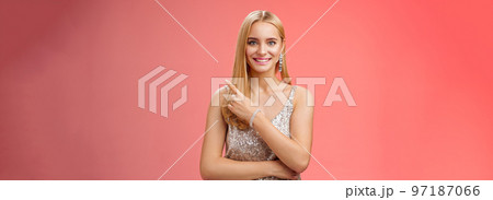 Confident attractive feminine glamour blond young woman in silver glittering dress smiling broadly pointing up discuss recent news promo sales standing friendly grinning camera red background 97187066