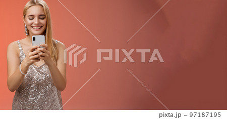 Delighted tender glamour blond woman in silver stylish glittering dress brilliand earrings holding smartphone taking photo friend capture moment celebration nightclub, red background smiling 97187195