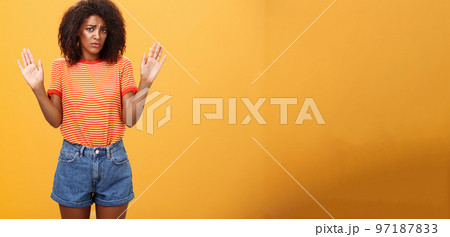 Silly insecure and sad dark-skinned female model in trendy striped t-shirt and shorts raising arms in surrender frowning being uninvolved and unaware standing clueless over orange background Silly insecure and sad dark-skinned female model in trendy striped t-shirt and shorts raising arms in surrender frowning being uninvolved and unaware standing clueless over orange background 97187833