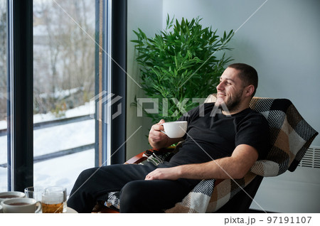 Young man enjoying weekends inside contemporary barn house. Happy male tourist sitting on chair, holding cup of tea, enjoying view of mountain landscape through panoramic windows in new cottage. Young man enjoying weekends inside contemporary barn house. Happy male tourist sitting on chair, holding cup of tea, enjoying view of mountain landscape through panoramic windows in new cottage. 97191107