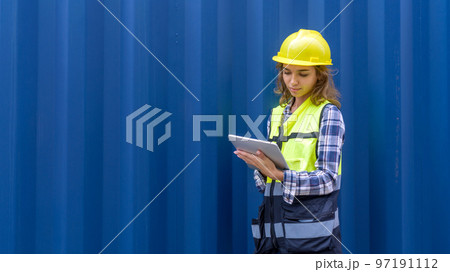 Young caucasian woman with safety vest and yellow hardhat checking shipping schedule on tablet computer, planning for next shipment. A large cargo container is in the background Young caucasian woman with safety vest and yellow hardhat checking shipping schedule on tablet computer, planning for next shipment. A large cargo container is in the background 97191112