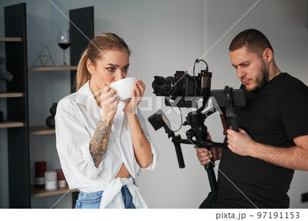 Videographer man shooting footage with female model, using camera mounted on gimbal stabilizer equipment indoors. Young woman with tattoos on hands posing and drinking hot tea. Videographer man shooting footage with female model, using camera mounted on gimbal stabilizer equipment indoors. Young woman with tattoos on hands posing and drinking hot tea. 97191153