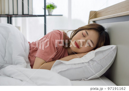 Young Asian woman in pink pajamas sleeping in bed leaning on pillows and covering white duvet. The atmosphere of the bedroom in the late afternoon, sunlight shining through the curtains and bookshelf. Young Asian woman in pink pajamas sleeping in bed leaning on pillows and covering white duvet. The atmosphere of the bedroom in the late afternoon, sunlight shining through the curtains and bookshelf. 97191162