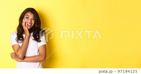 Portrait of attractive african-american girl, looking amazed and smiling, standing over yellow background in casual white t-shirt Portrait of attractive african-american girl, looking amazed and smiling, standing over yellow background in casual white t-shirt 97194125