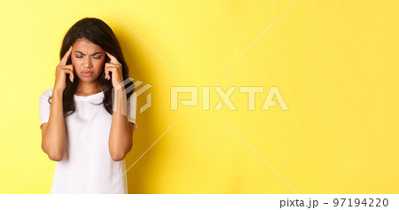 Portrait of young african-american female model feeling sick, frowning and touching head, complaining on headache, standing over yellow background Portrait of young african-american female model feeling sick, frowning and touching head, complaining on headache, standing over yellow background 97194220