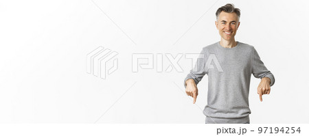 Handsome and confident middle-aged man in grey sweater, pointing fingers down and smiling, showing good deal promo, standing over white background Handsome and confident middle-aged man in grey sweater, pointing fingers down and smiling, showing good deal promo, standing over white background 97194254