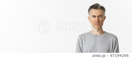 Close-up of skeptical middle-aged man, frowning and looking doubtful at camera, standing over white background in gray sweater 97194266