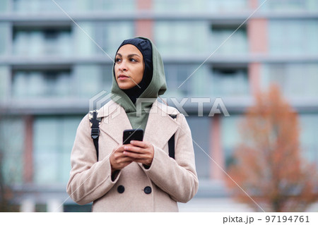 Portrait of young muslim woman in coat, standing outdoor in city, scrolling smartphone. Portrait of young muslim woman in coat, standing outdoor in city, scrolling smartphone. 97194761