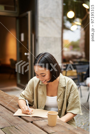 Vertical shot of asian girl student, writing smth in notebook, making notes, smiling while doing homework outdoors from cafe 97195315