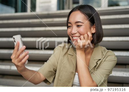 Portrait of happy smiling asian girl takes selfie on mobile phone. Young korean woman video chat on smartphone while sitting on stairs near building Portrait of happy smiling asian girl takes selfie on mobile phone. Young korean woman video chat on smartphone while sitting on stairs near building 97195316