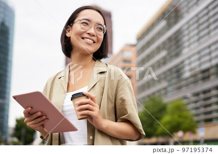 Portrait of happy young woman in glasses, standing on street with cup of coffee and tablet 97195374