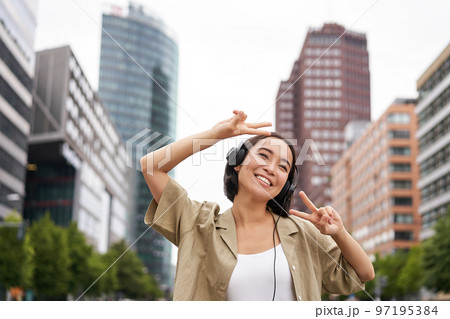 Happy asian woman in headphones, listening music and dancing on street of city centre, smiling with hands up 97195384