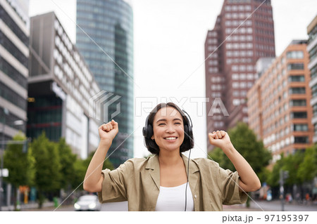 Portrait of smiling asian woman dancing, triumphing, feeling happy while listening music in city, posing on street near skyscrappers Portrait of smiling asian woman dancing, triumphing, feeling happy while listening music in city, posing on street near skyscrappers 97195397