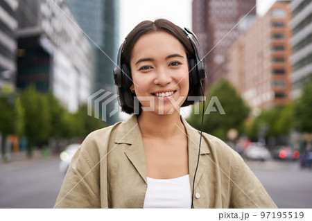 Portrait of smiling asian woman in headphones, standing in city centre on street, looking happy, listening to music Portrait of smiling asian woman in headphones, standing in city centre on street, looking happy, listening to music 97195519