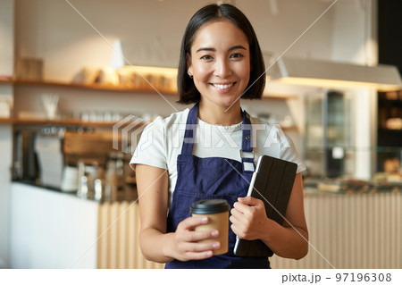 Smiling barista, cafe waitress giving out takeaway coffee order, prepared cappuccino for takeout, standing in blue apron with tablet 97196308
