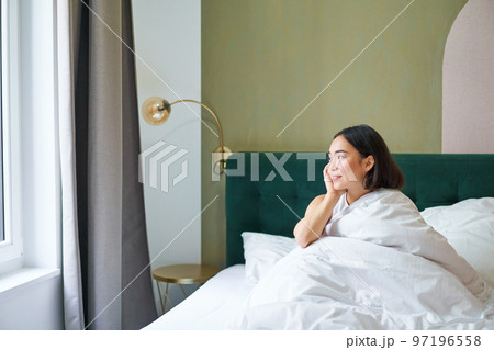 Beautiful sleepy asian girl wakes up in her bed with cozy white blankets, looking outside window with pleased smile. Lifestyle concept Beautiful sleepy asian girl wakes up in her bed with cozy white blankets, looking outside window with pleased smile. Lifestyle concept 97196558