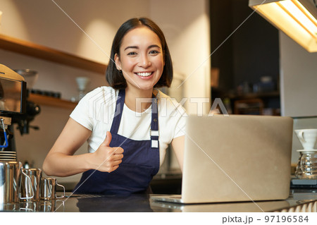 Portrait of asian girl in apron, barista in coffee shop, shows thumbs up, approve or recommend smth, working in cafe, smiling happy 97196584