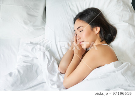 Top view image of asian woman sleeping alone in king size bed on white pillows. Young girl lying in her bedroom with eyes closed, morning sunlight shines in room Top view image of asian woman sleeping alone in king size bed on white pillows. Young girl lying in her bedroom with eyes closed, morning sunlight shines in room 97196619