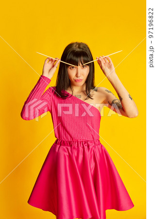 Young emotive girl in bright pink dress making brows with chopsticks isolated on vivid yellow background. Concept of youth, beauty, fashion, lifestyle, emotions 97196822