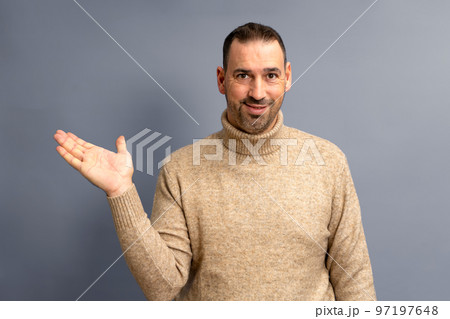Hispanic man with beard and brown turtleneck sweater in friendly attitude offering something on the palm of his hand, isolated on gray background. Hispanic man with beard and brown turtleneck sweater in friendly attitude offering something on the palm of his hand, isolated on gray background. 97197648